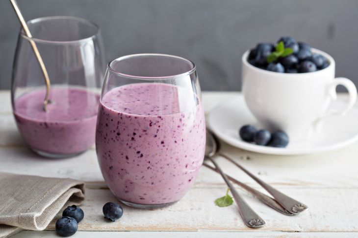 Two glasses of thick blueberry almond butter shake on a light wooden surface, with fresh blueberries scattered nearby and a white teacup filled with blueberries in the background. A napkin and spoons are placed beside the drinks.