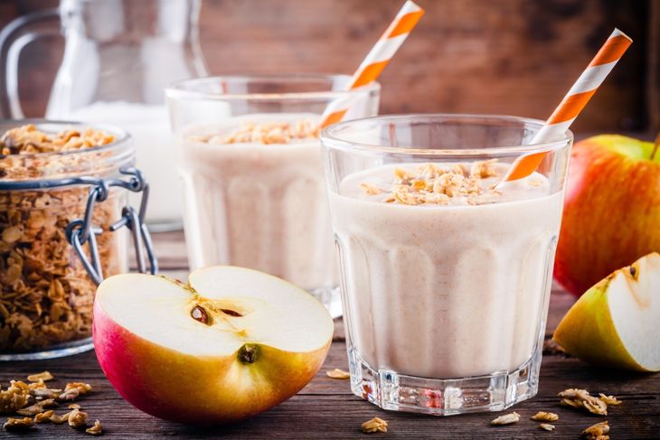 Two glasses of creamy apple crisp smoothie topped with granola and served with orange-striped straws, surrounded by fresh apple slices, a jar of oats, and a rustic wooden background, evoking cozy autumn flavors.