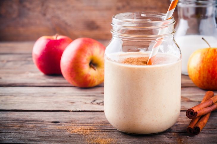 A mason jar filled with a light brown smoothie topped with cinnamon, with a striped straw, surrounded by apples, cinnamon sticks, and a jar of milk—visually representing a spiced apple oat blend.