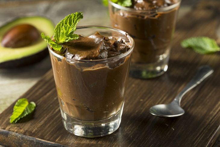 A glass filled with thick chocolate mousse garnished with a sprig of fresh mint, with another glass in the background and half an avocado nearby—representing an avocado-based chocolate smoothie.