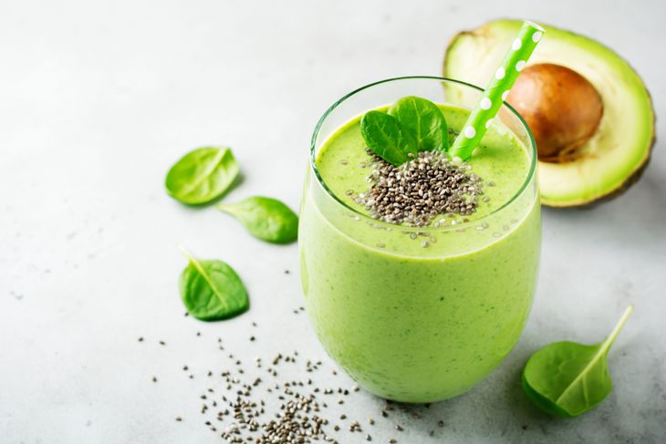 A glass of green avocado-spinach smoothie topped with chia seeds and a spinach leaf, surrounded by baby spinach leaves and a halved avocado with the pit visible. A green polka-dotted straw is placed in the glass.