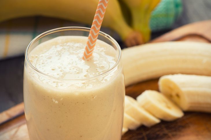 A creamy banana smoothie in a glass with an orange-striped straw sits in front of a peeled banana and sliced banana pieces on a wooden board. The background includes a whole banana and a patterned towel.