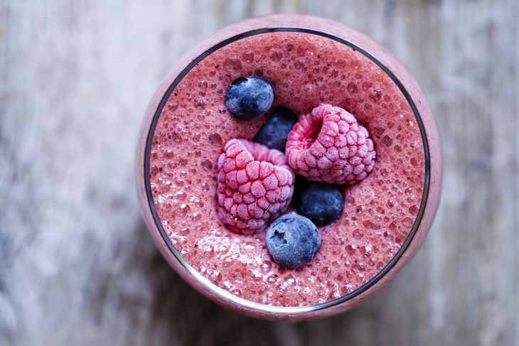 A close-up view of a blended berry smoothie topped with frozen raspberries and blueberries, served in a clear glass on a rustic wooden surface.