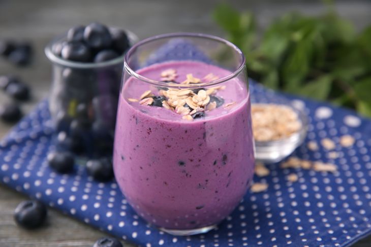 A thick blueberry oat smoothie in a clear glass topped with oats and whole blueberries, set on a dark blue polka-dotted cloth with scattered oats and berries nearby, evoking a hearty, wholesome breakfast.