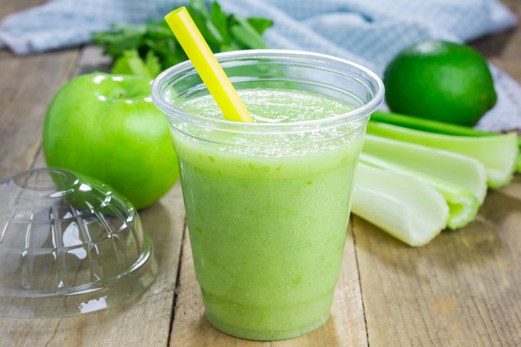 A plastic cup filled with a bright green smoothie and a yellow straw, surrounded by fresh celery stalks, a green apple, a lime, and leafy greens on a wooden surface. A dome lid sits beside the cup.