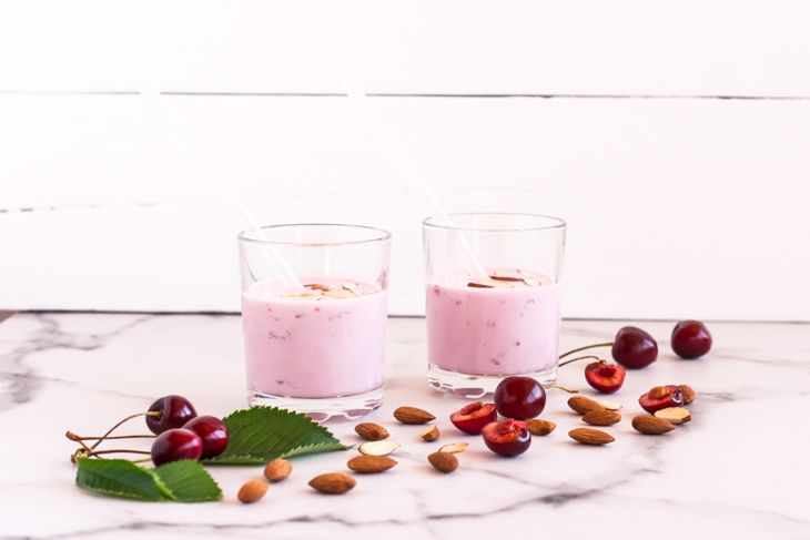 Two glasses of pink smoothie with white straws, topped with almond slices, surrounded by whole cherries, halved cherries, almonds, and green leaves on a marble surface.