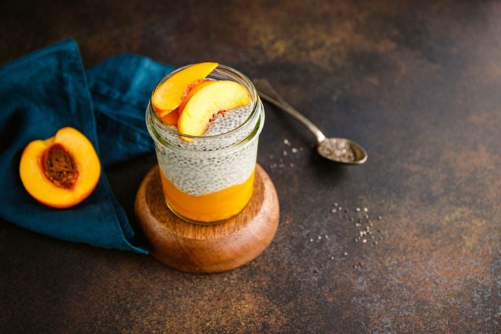 A jar filled with layered chia peach overnight smoothie sits on a wooden coaster, topped with fresh peach slices. A spoonful of chia seeds and a halved peach rest nearby, highlighting the main ingredients.