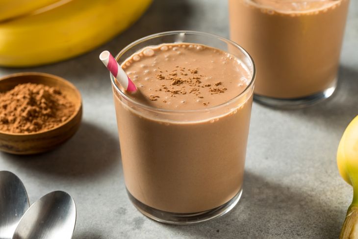 A glass of chocolate banana protein shake topped with a light dusting of cocoa powder, with a striped straw, surrounded by bananas, a bowl of cocoa powder, and metal spoons on a gray surface.