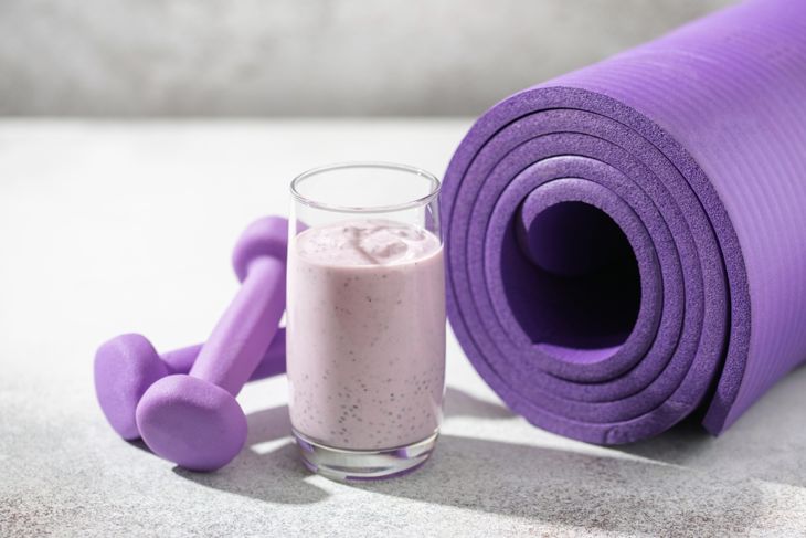 A glass of thick chocolate berry protein smoothie sits in front of a rolled purple yoga mat and a pair of matching purple dumbbells, suggesting a post-workout recovery setting.