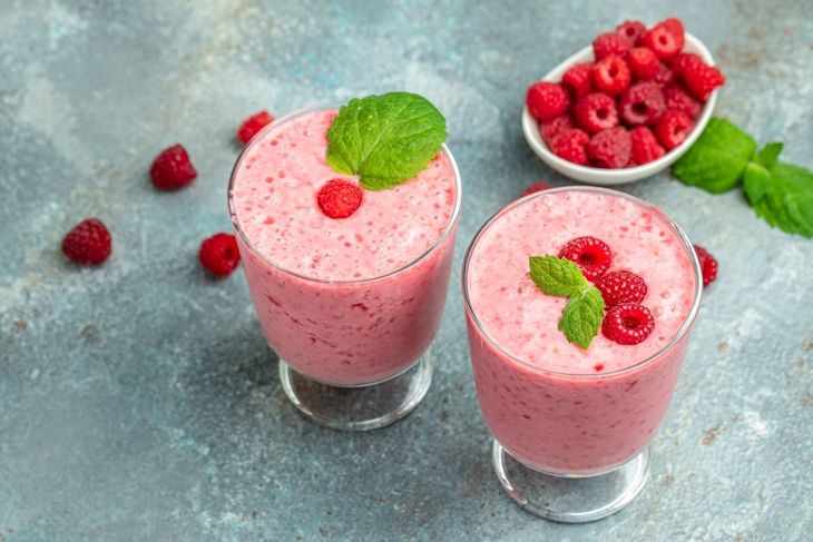 Two glasses filled with a pink raspberry smoothie, garnished with fresh raspberries and mint leaves, surrounded by scattered raspberries and a small bowl of berries—illustrating a coconut and raspberry blend.