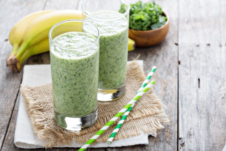 Two tall glasses filled with a thick green smoothie sit on a rustic wooden table, surrounded by bananas, kale, and green-striped straws.