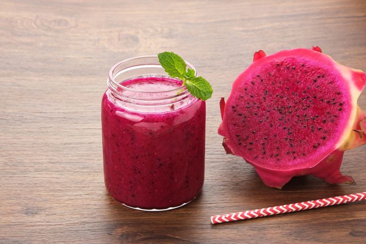 A magenta-colored smoothie in a clear mason jar, topped with a mint sprig, placed next to a halved dragon fruit and a red-striped straw on a wooden surface.