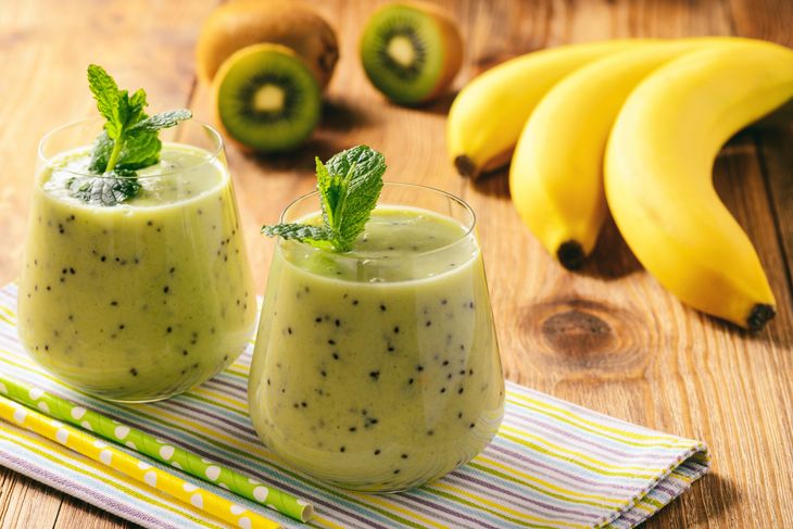 Two glasses of green kiwi smoothie garnished with fresh mint, placed on a striped cloth with yellow bananas and halved kiwis in the background. The smoothie contains visible kiwi seeds throughout.