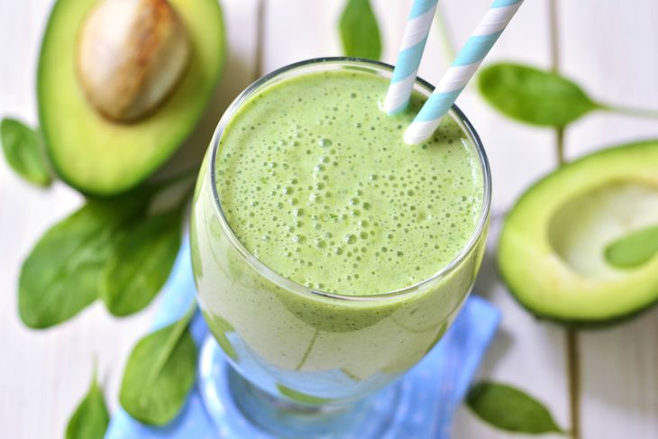 A tall glass filled with a light green smoothie sits on a blue napkin, surrounded by spinach leaves and halved avocados, with two striped paper straws placed in the drink.