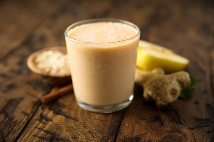 A creamy light-orange smoothie in a clear glass, placed on a rustic wooden surface with fresh ginger, sliced banana, oats, and a cinnamon stick in the background.