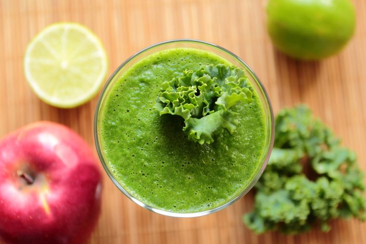 A glass of bright green kale smoothie topped with a curly kale leaf, surrounded by a red apple, a halved lemon, a green lime, and fresh kale on a bamboo surface.