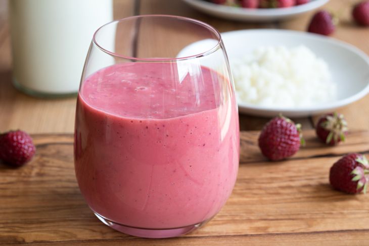 A glass of thick, pink berry smoothie made with kefir sits on a wooden surface, surrounded by fresh strawberries, with a white plate of kefir grains and a glass jar of milk in the background.