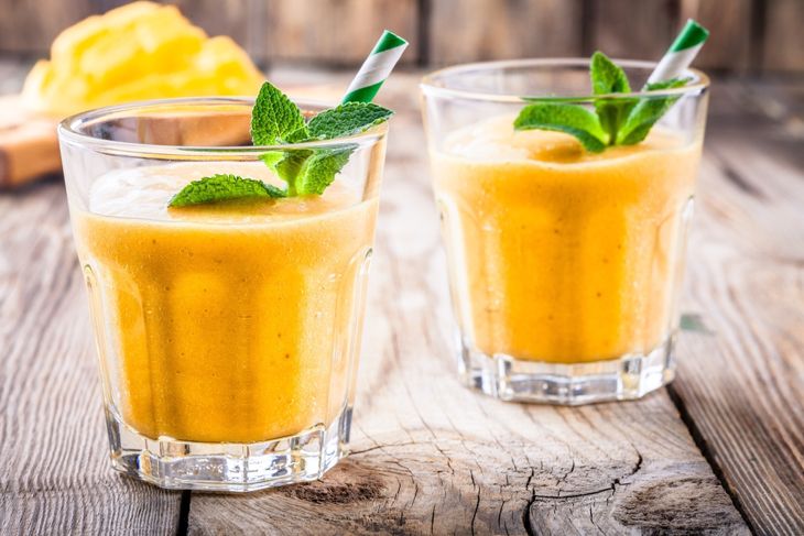 Two glasses of vibrant yellow mango pineapple smoothie topped with fresh mint leaves and striped straws sit on a rustic wooden table, offering a bright and tropical refreshment.