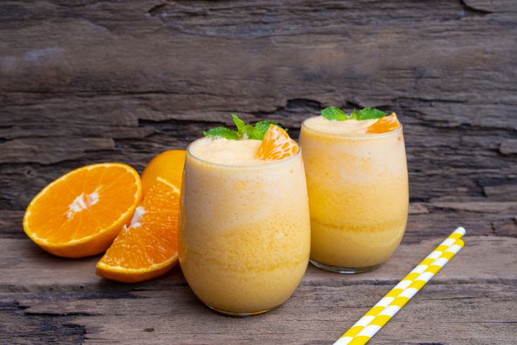 Two glasses of orange smoothie are topped with fresh orange segments and mint leaves, sitting on a rustic wooden surface with halved oranges and striped yellow straws nearby. The image reflects the creamy, citrus-focused ingredients of an orange creamsicle smoothie.