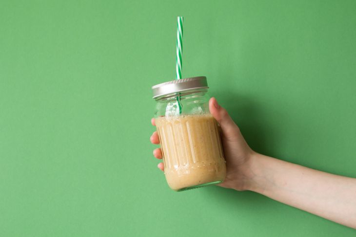 A hand holds a mason jar filled with a light brown smoothie, topped with a metal lid and green-striped straw, set against a solid green background. The smoothie reflects the peanut butter and fruit blend of a PB&J-inspired drink.