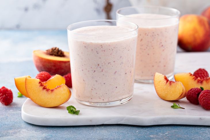 Two glasses of light pink smoothie on a white serving board, surrounded by fresh peach slices, whole raspberries, and a halved peach in the background.