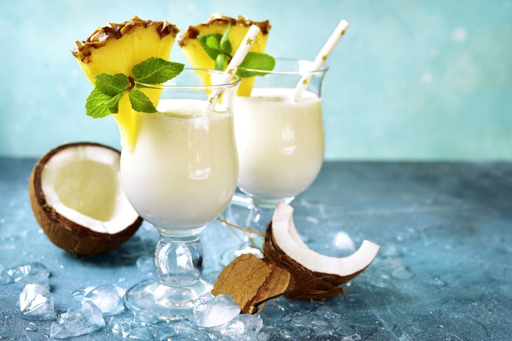 Two glasses of creamy white piña colada smoothies are garnished with pineapple wedges, mint sprigs, and paper straws. Pieces of fresh coconut and ice cubes are scattered on the surface nearby.