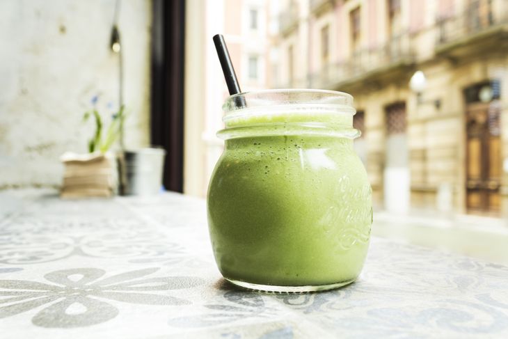 A mason jar filled with a green smoothie and a black straw, sitting on a patterned table near a window with an urban street view in the background.