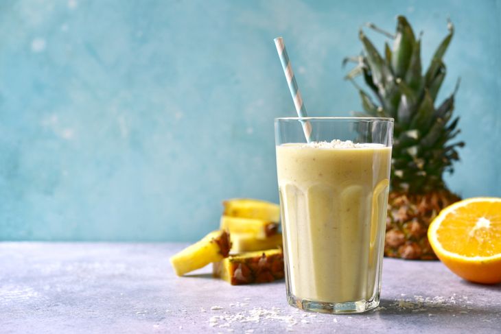 A tall glass of pale yellow pineapple orange smoothie with a paper straw, surrounded by pineapple slices, a halved orange, and a whole pineapple in the background.