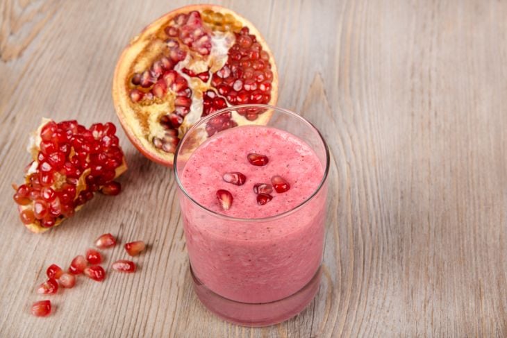 A glass of bright pink pomegranate berry smoothie topped with a few scattered pomegranate seeds sits on a wooden surface next to a halved pomegranate and loose arils, capturing the rich color and refreshing appeal of this antioxidant-packed drink.