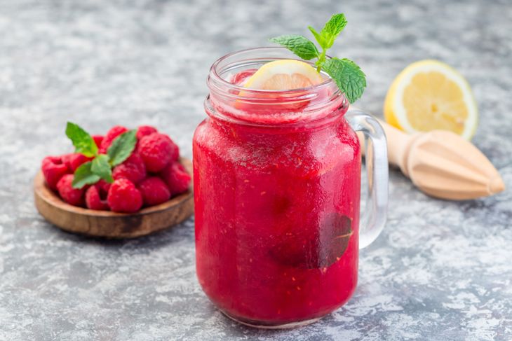 A mason jar filled with a bright raspberry lemonade smoothie, garnished with a lemon slice and mint sprig, with fresh raspberries and a halved lemon nearby.
