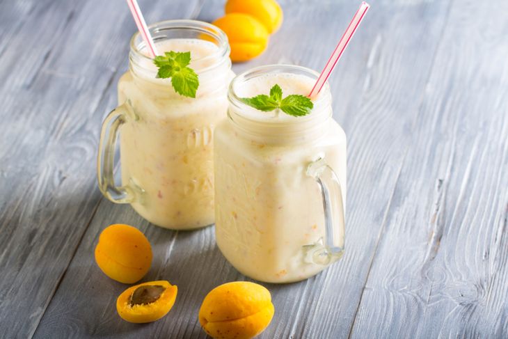 Two mason jars filled with creamy apricot-colored smoothies, each garnished with a sprig of mint and a striped straw. Fresh apricots, whole and halved, are scattered on the wooden surface around the drinks.