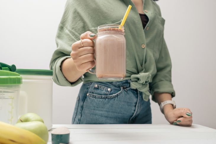 A person in a green shirt and jeans holds a mason jar filled with a chocolate smoothie topped with cocoa powder and a yellow straw. Nearby are a protein scoop, bananas, green apples, and containers on a white table.