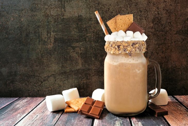 A chocolate smoothie served in a glass mug topped with mini marshmallows, graham cracker pieces, and chocolate squares, styled to reflect the ingredients of a s’mores-inspired blend featuring cocoa, banana, and oat milk.