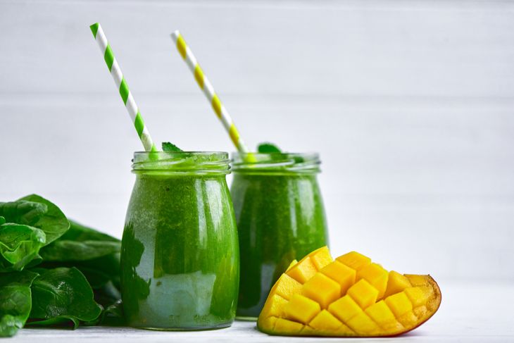 Two glass jars filled with a green smoothie, each topped with a mint sprig and striped paper straw, alongside a halved, cubed mango and fresh spinach leaves on a white surface.