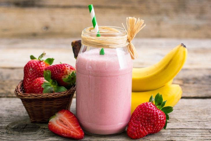 A glass jar filled with a creamy pink strawberry banana smoothie, topped with a green-striped straw and tied with rustic twine, sits surrounded by fresh strawberries and bananas on a wooden table.