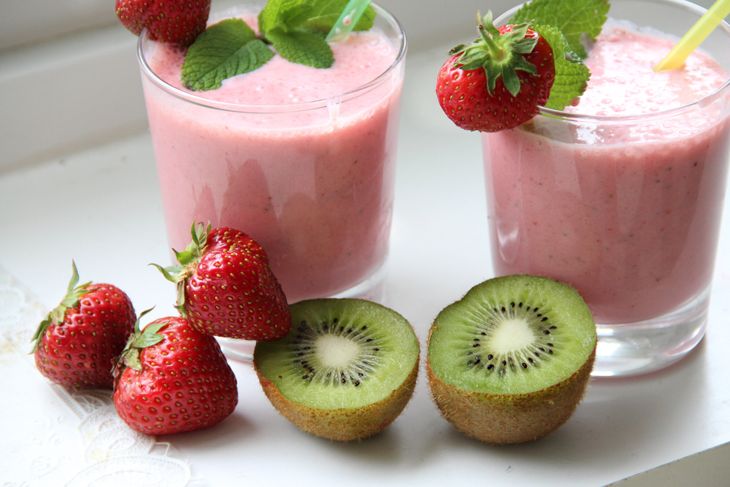 Two glasses of pink strawberry kiwi smoothie topped with mint leaves and whole strawberries, surrounded by fresh strawberries and a halved kiwi fruit on a white surface.