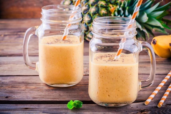 Two mason jars filled with a creamy orange smoothie topped with chia seeds, each with a striped straw, surrounded by tropical fruits like pineapple and banana, representing a mango-pineapple blend with coconut milk and chia.