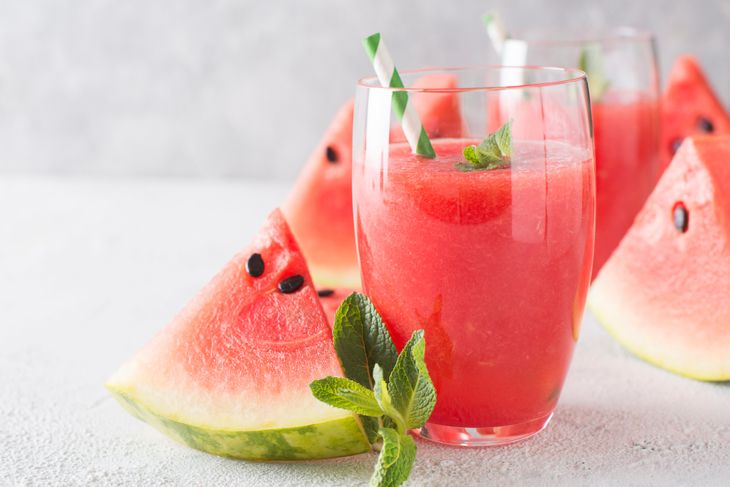 Glasses of vibrant pink watermelon mint smoothie garnished with fresh mint and green-striped straws, surrounded by juicy watermelon slices on a light stone surface—summer refreshment at its best.