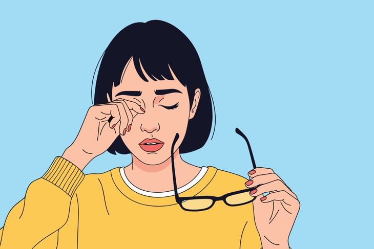 An illustration shows a woman rubbing one eye while holding a pair of glasses in her other hand against a plain blue background.