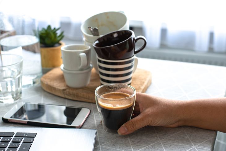 A young female freelancer working from home holding a cup of coffee with many cups in the background drinks too much coffee caffeine addiction anxious and crazy in maniac bad lifestyle concept