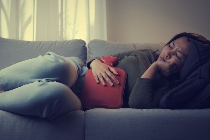Asian woman has sick heavy menstrual pain applying red hot water bag on her stomach to relieve the pain on the living room sofa