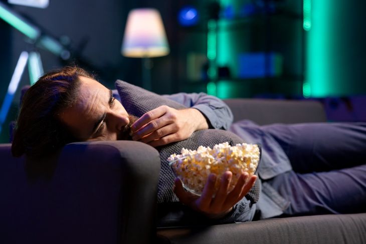 Man late at night lounged on couch, eating popcorn and watching movies on TV before going to sleep. Person relaxing at home, enjoying snacks and film trying to fall asleep