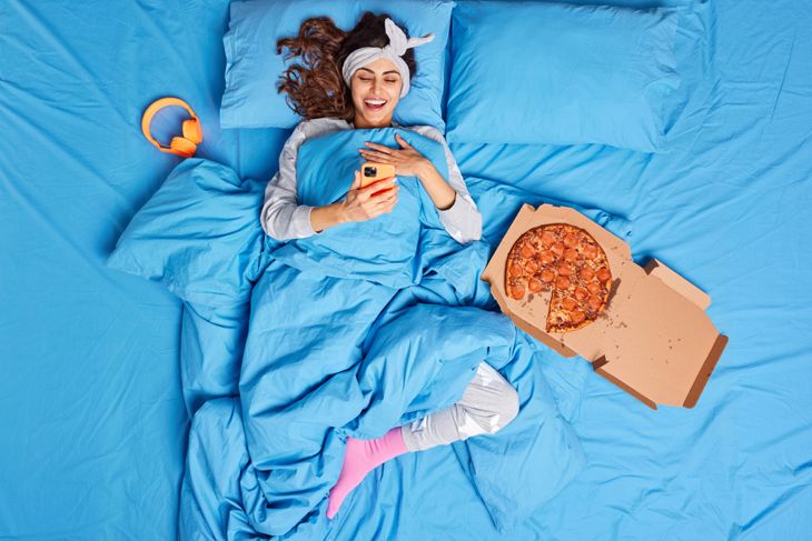 A woman lies in bed under bright blue sheets, smiling at her phone while wearing pajamas, a headband, and pink socks. A partially eaten pepperoni pizza sits in an open box beside her, suggesting late-night snacking and screen time.