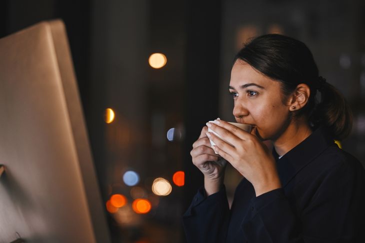 A woman sits at a computer at night, holding a white mug near her face. The screen glows in front of her, and blurred city lights are visible through the window behind her—suggesting late-evening caffeine use.