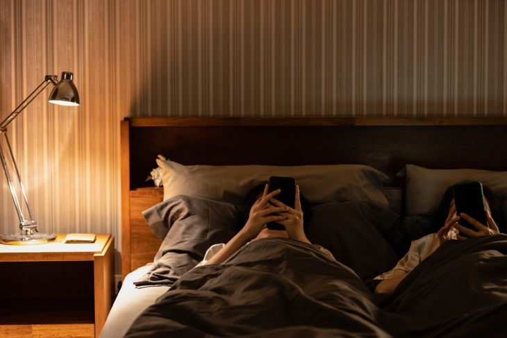 Certainly — here’s revised alt text that includes a nod to the “digital sunset” concept while maintaining editorial clarity and tone: Two people lie in bed in a dimly lit room, each looking at a smartphone. A bedside lamp glows nearby. The scene contrasts with a true “digital sunset” routine, where screens are put away before sleep.