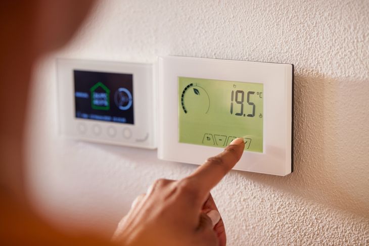 Rear view of multi ethnic woman pressing a button of a wall attached house thermostat with digital display showing the temperature. 