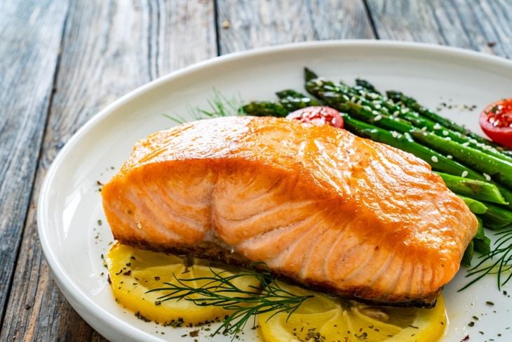 Fried salmon steak with cooked green asparagus, cherry tomatoes and lemon slices served on white plate on wooden table
