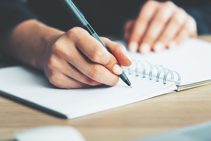 Close up of woman's hands writing in spiral notepad placed on wooden desktop with various items