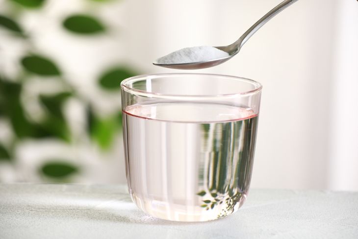 Spoon with baking soda over glass of water at white table against blurred background, closeup