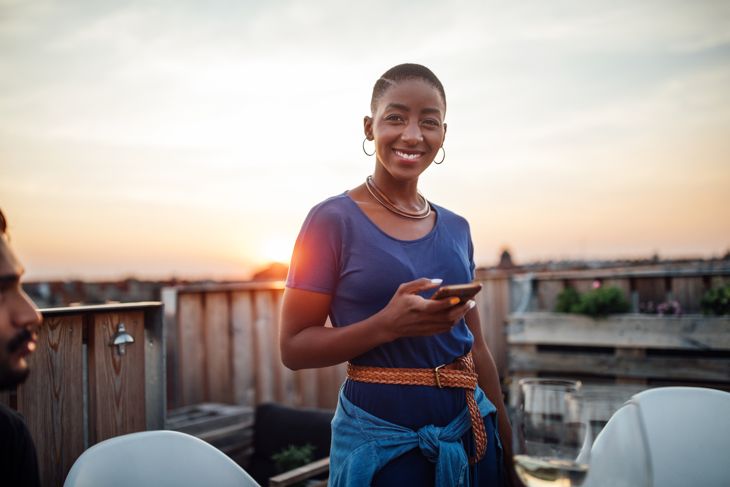 Beautiful young african woman with bald head standing on roof holding her mobile phone. Female at rooftop party with phone at sunset.